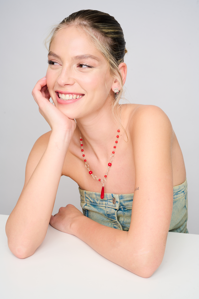 תמונה של שרשרת קלילה אך מושכת עין, עם חרוזי אלמוג סינתטי אדומים בגדלים שונים, וטבעות כסף עדינות. התליון המרכזי בצורת טיפה מדגיש את קו המחשוף ומושך את העין. הלוק הכללי משדר חופש, אנרגיה נשית ותחושת נועזות עדינה.