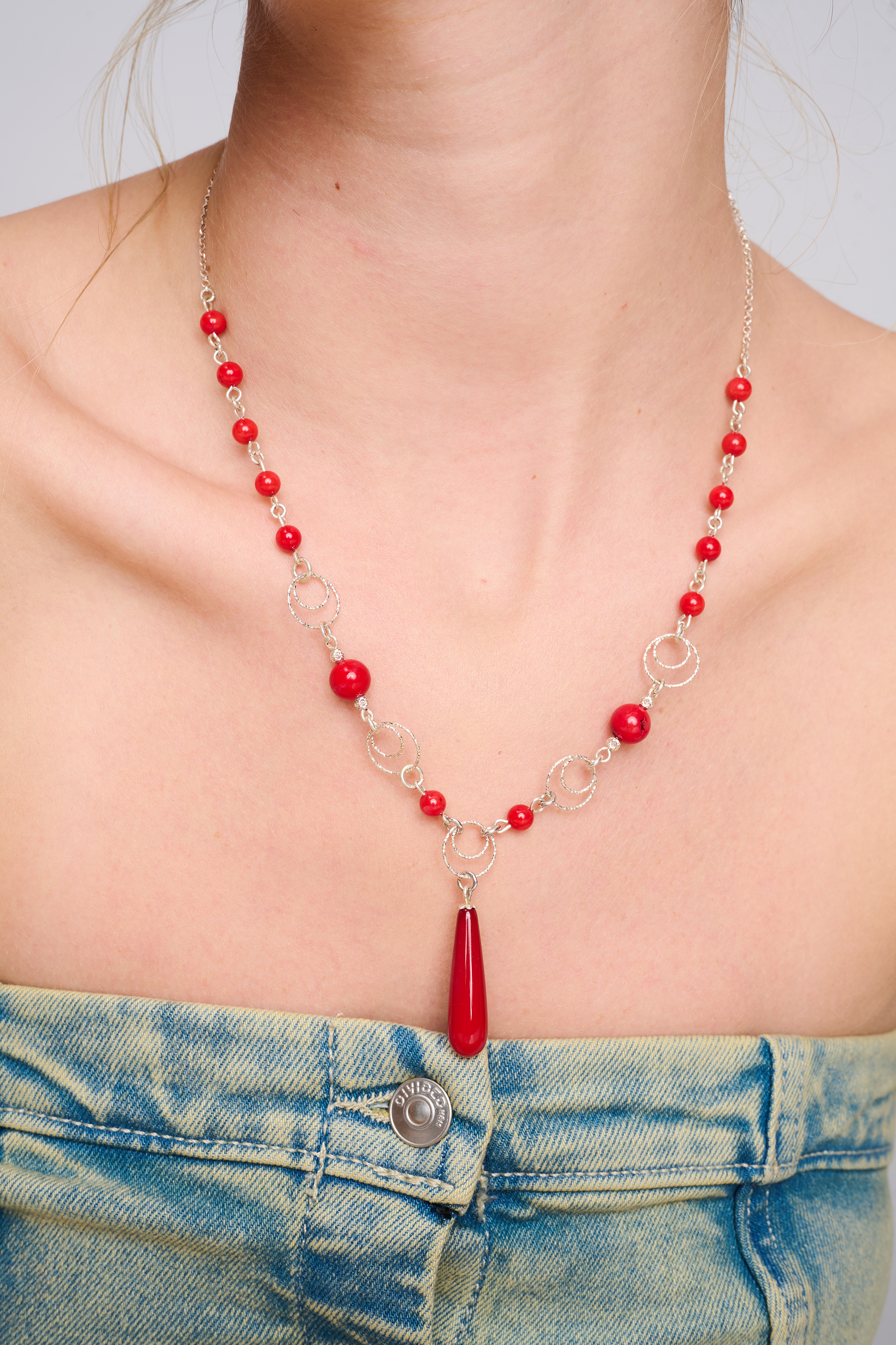 תמונה של שרשרת עדינה מכסף עם חרוזים באדום בוהק, עשויים מאבן אלמוג סינתטי. במרכז תלוי תליון בטיפה ארוכה בגוון תואם. השרשרת משלבת בין חרוזים עגולים קטנים וגדולים לבין טבעות כסף דקורטיביות. מוצגת על צוואר חשוף של דוגמנית לבושה בג'ינס, באווירה קלילה ונשית.